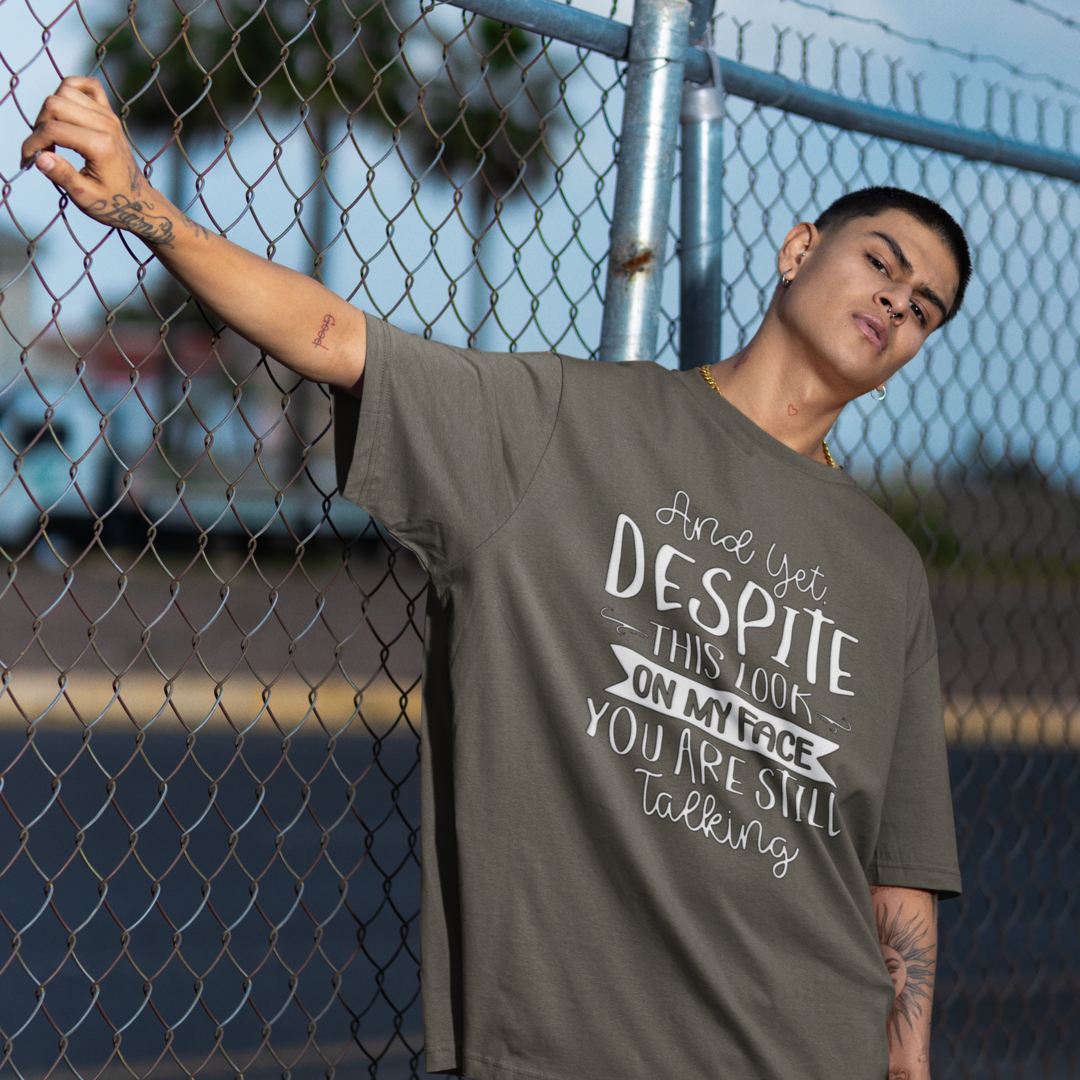 A Man Leaning Against a Chain Link Fence, Wearing A Crew Neck T-Shirt with Text - "And Yet, Despite This Look on My Face, You&